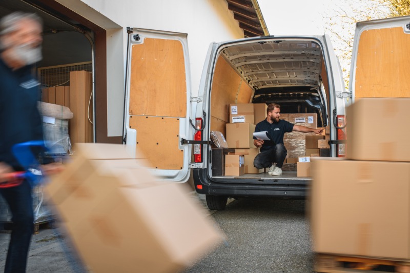 A delivery worker organising cardboard boxes inside a van while another worker carries a package.
