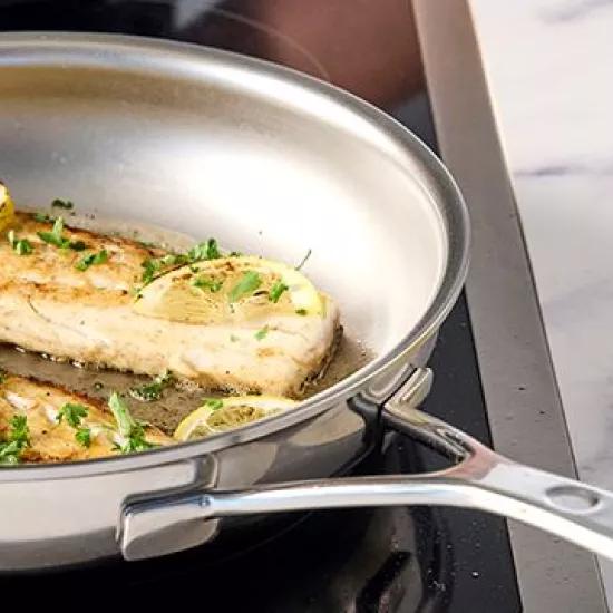 Een roestvrijstalen pan met gebakken visfilets, gegarneerd met citroenschijfjes en peterselie, op een inductiekookplaat. De pan heeft een antifkleeftextuur als PFAS-alternatief.