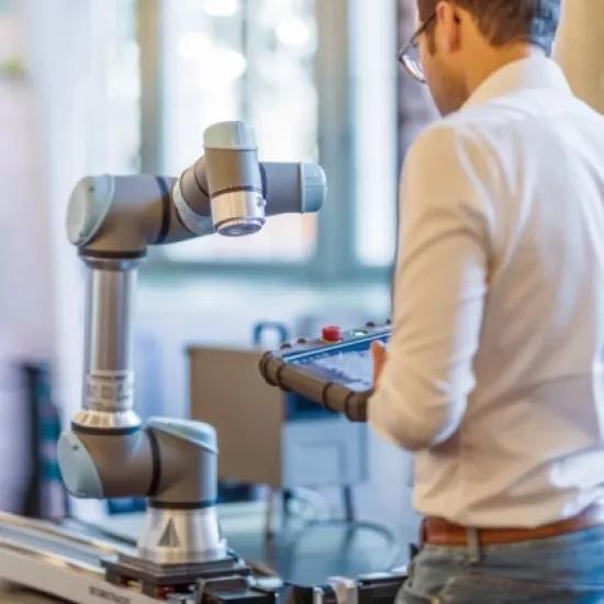 A man operates a collaborative robot arm (cobot) using a control panel in an industrial environment.