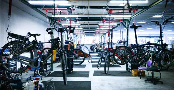 Een fietsrevisiewerkplaats waar meerdere tweedehands elektrische leasefietsen aan een bovenrailsysteem hangen voor onderhoud. Technici van maatwerkbedrijf Ecoso repareren en optimaliseren de fietsonderdelen om hun levensduur te verlengen. Dit initiatief ondersteunt productlevensduurverlenging en de circulaire economie door gebruikte fietsen een tweede leven te geven via duurzame revisie.