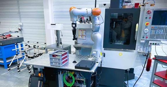 A mobile cobot assistant on a wheeled platform in an industrial environment, working next to a CNC machine. The cobot, branded KUKA, is designed to automate repetitive tasks in manufacturing. The setup includes cables, a control panel, and storage bins for small parts.