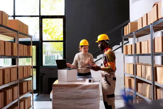 Deux employés d'entrepôt portant des casques discutent des processus de travail près d'un ordinateur portable, entourés d'étagères de boîtes, illustrant l'efficacité dans la réduction du Work-in-Progress (WIP).